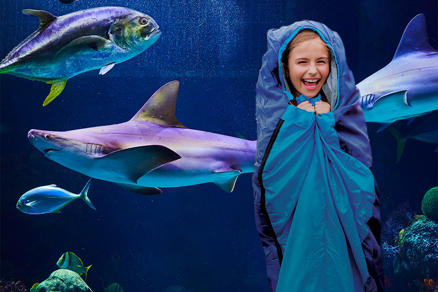A young girl wrapped in a blanket and sleeping bag laughing while standing in front of an aquarium with sharks and fish.
